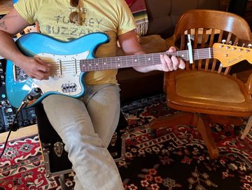 Person playing blue electric guitar while sitting indoors.