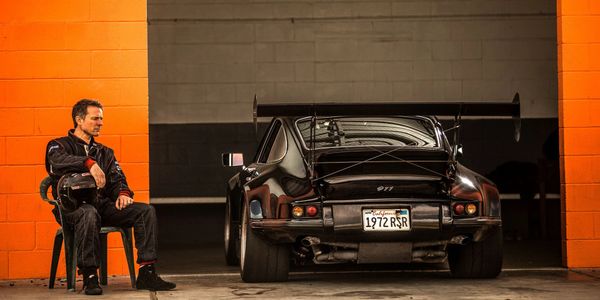 A man in a racing suit and helmet sitting by his black Porsche