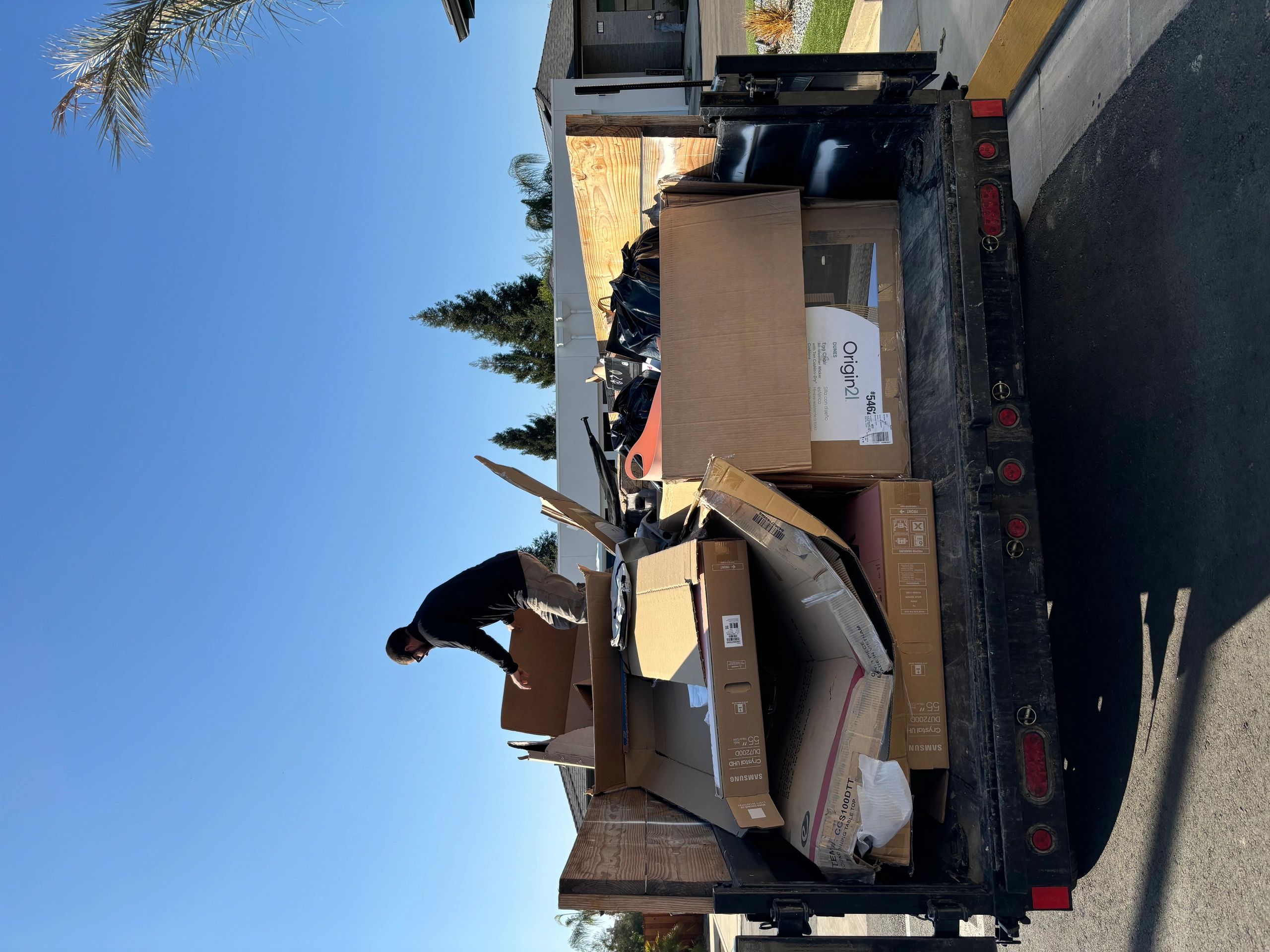 Man organizing large cardboard boxes and black bags on a truck bed under clear blue sky.