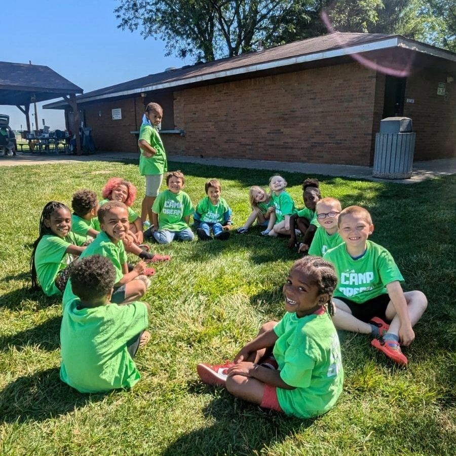 Children in green shirts sitting in a circle on grass at camp.
