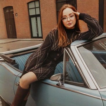 Young woman in glasses leaning on a vintage blue car wearing a stylish outfit and boots.
