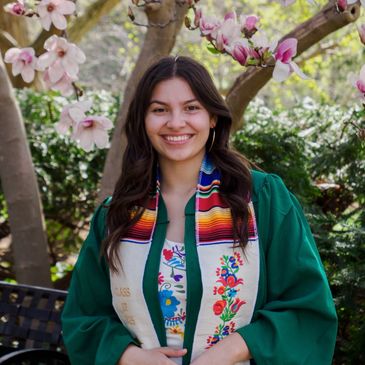 Smiling graduate in green gown and colorful stole stands among blooming magnolia trees.