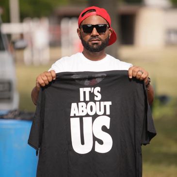 Man holding a black t-shirt with the text 'IT'S ABOUT US'.