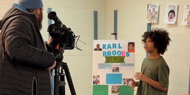 A young boy presents a project about Karl Brooks while being filmed.