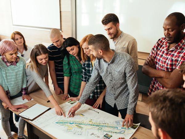 A diverse group of young adults gathered around a table, pointing at a large plan.