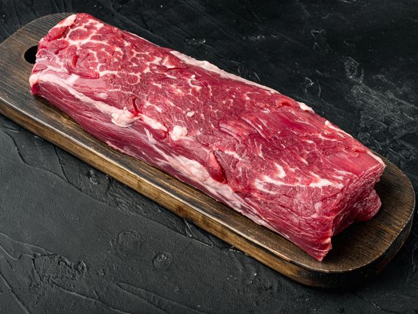 A large, rectangular cut of raw beef tenderloin with visible marbling rests on a dark wooden board.