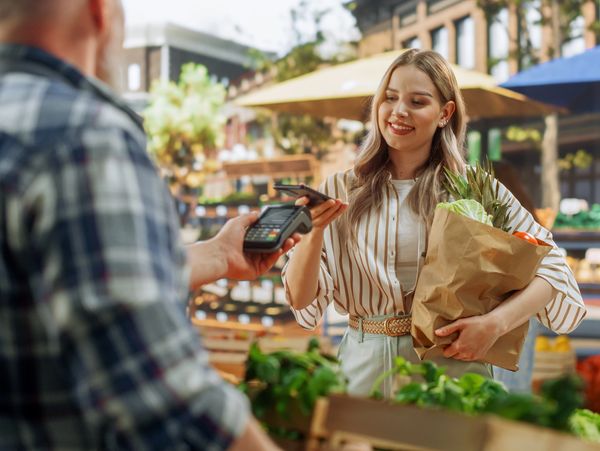 Smiling woman paying with card for a bag of fresh produce at a farmers market.