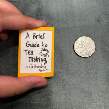 Hand holding a tiny book on tea making next to a coin.