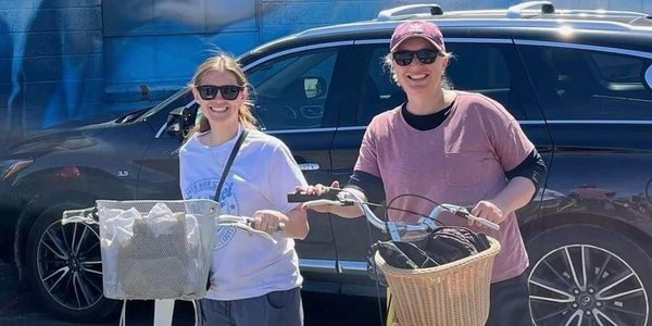 Two women smiling with bicycles in front of a black car.