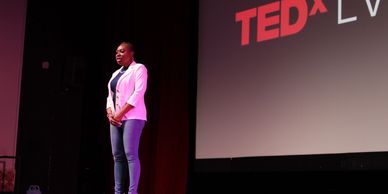A woman speaking on stage at a TEDx event, wearing a white blazer and jeans.