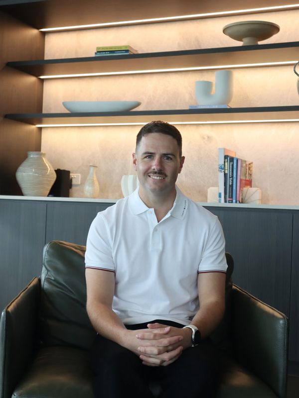 Man in white polo shirt sitting on a couch with a modern bookshelf behind him.