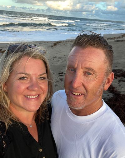 A smiling couple takes a selfie on a beach with waves and cloudy sky in the background.