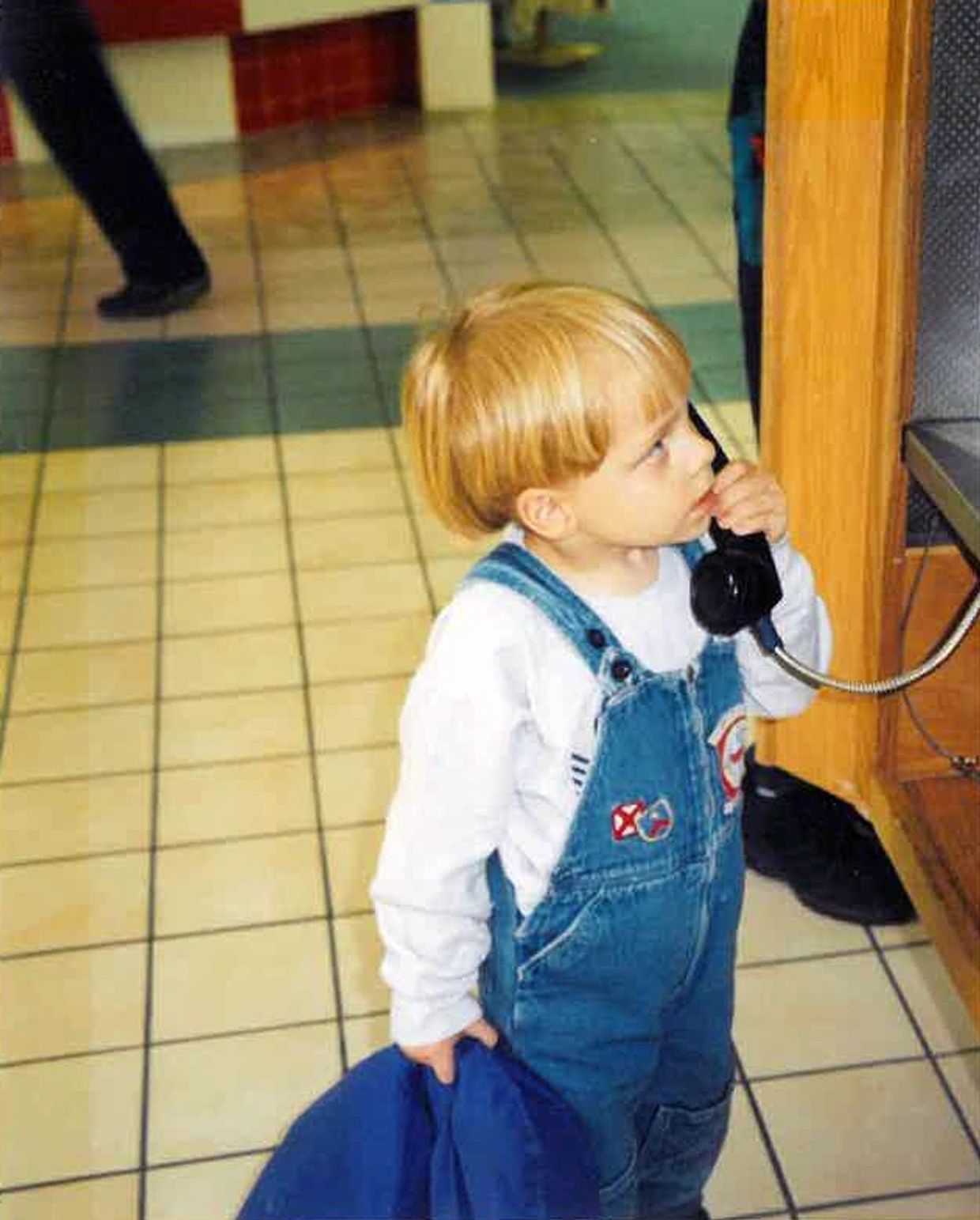 Young boy with blonde bowl cut and jean overalls speaking into a public payphone in a bustling mall.