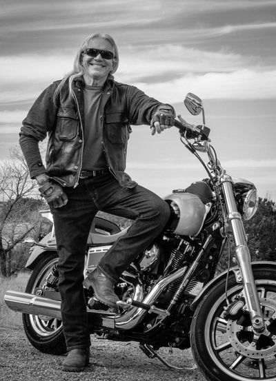 Smiling man in leather jacket posing with motorcycle outdoors.