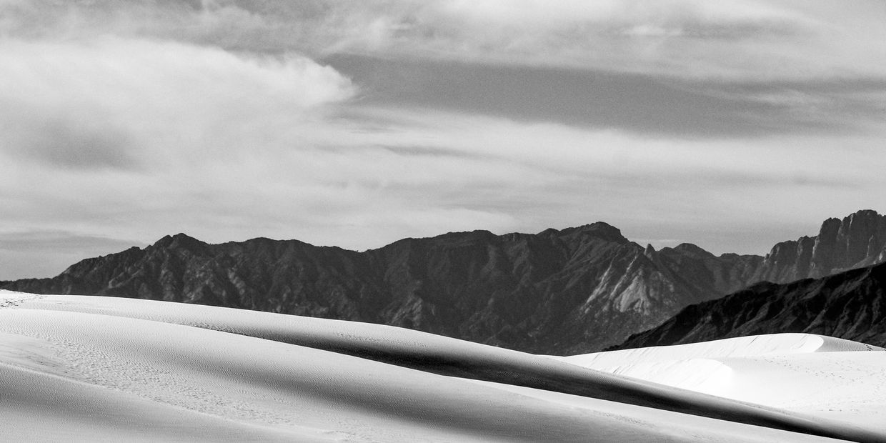 Black and white image of sand dunes with mountains in background.