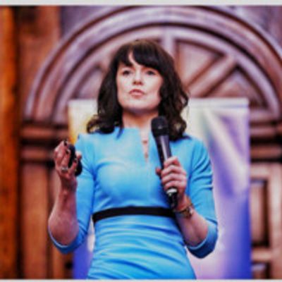 Prof Maura McAdam in a blue dress standing with a microphone in her left hand giving a keynote.