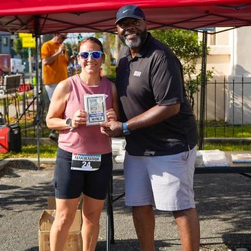 Two people posing with a winner plaque at an outdoor event.