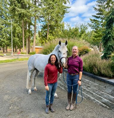 Two women standing with a white horse in an outdoor setting surrounded by greenery.