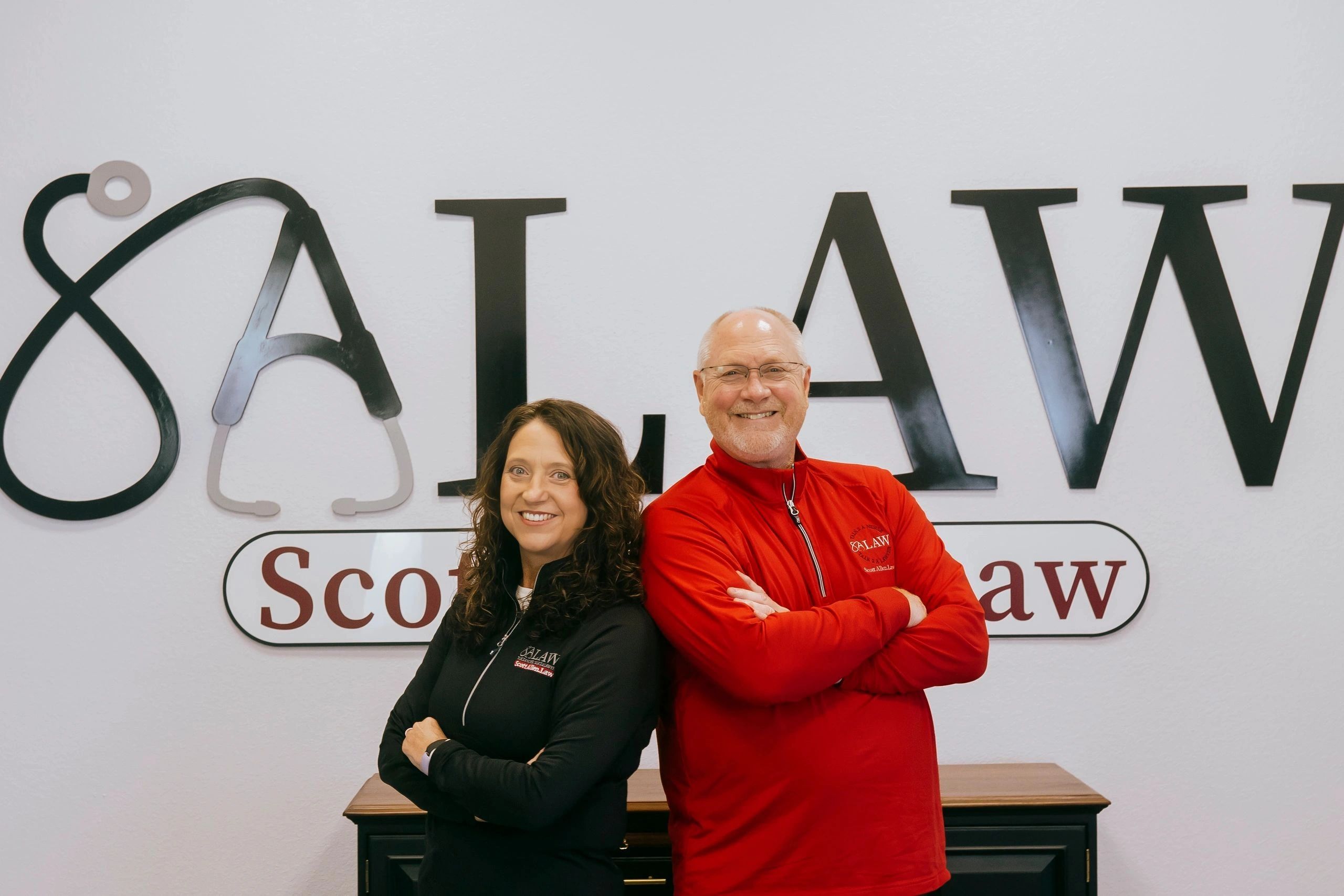 Image of Scott and Leslie Allen standing back-to-back in front of the Scott Allen.Law sign.