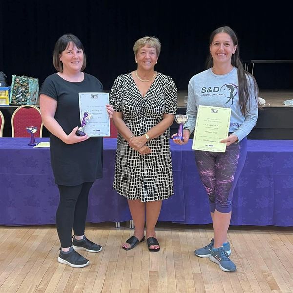Three women posing indoors, two holding certificates and medals.