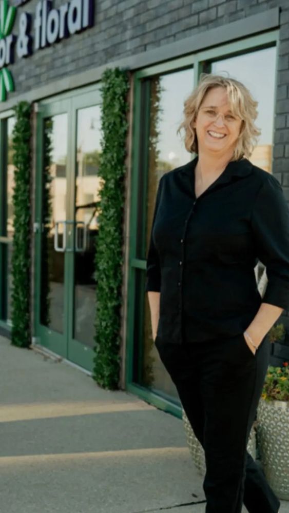 A smiling woman in black stands outside a floral store named Lazy Acres.