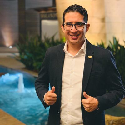 Man in glasses and suit giving thumbs up by a pool at night.