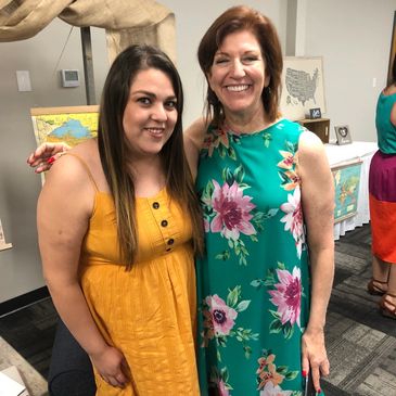 Two women smiling, one in a yellow dress and the other in a floral green dress.