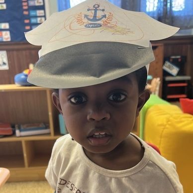 A young child wearing a paper captain's hat indoors.