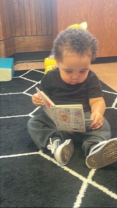 A toddler sitting on a rug, deeply focused on reading a book.