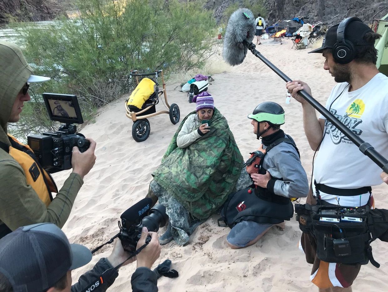 On a sandy area along the river, Cara sits wrapped in a blanket drinking hot tea, talking to Roy.