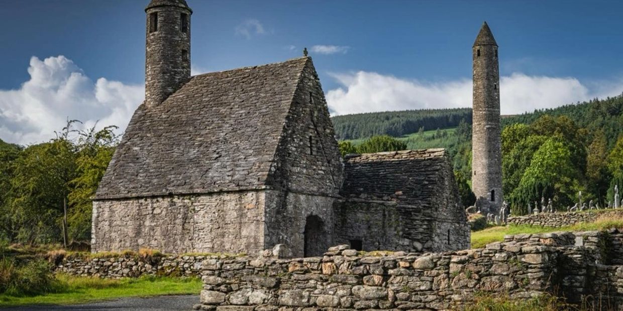 Ancient stone church and round tower in a lush green countryside under a blue sky.