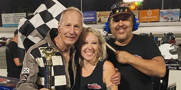 Three people celebrating at a race track, one holding a trophy and a checkered flag.