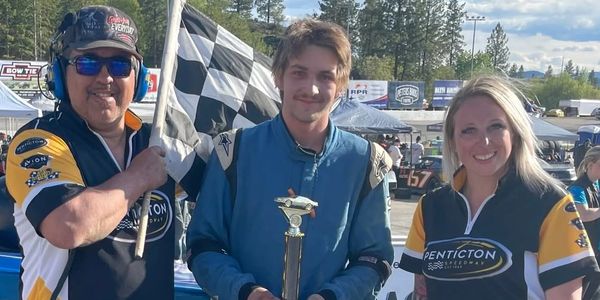 Race winner holding trophy with team members and checkered flag at racetrack.