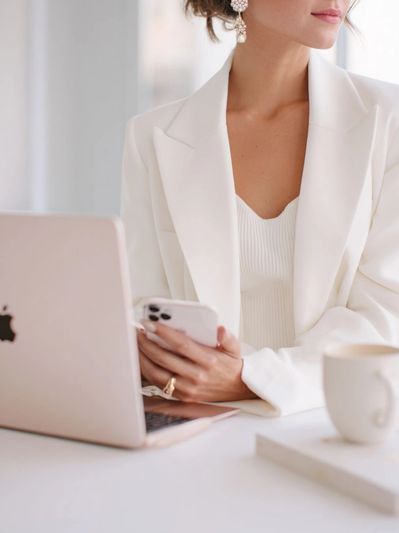 Woman in white blazer holding phone, working on a laptop.