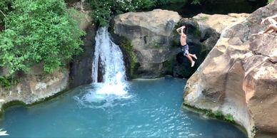 Boy jumping from a rocky cliff into a pond in front of a waterfall.
