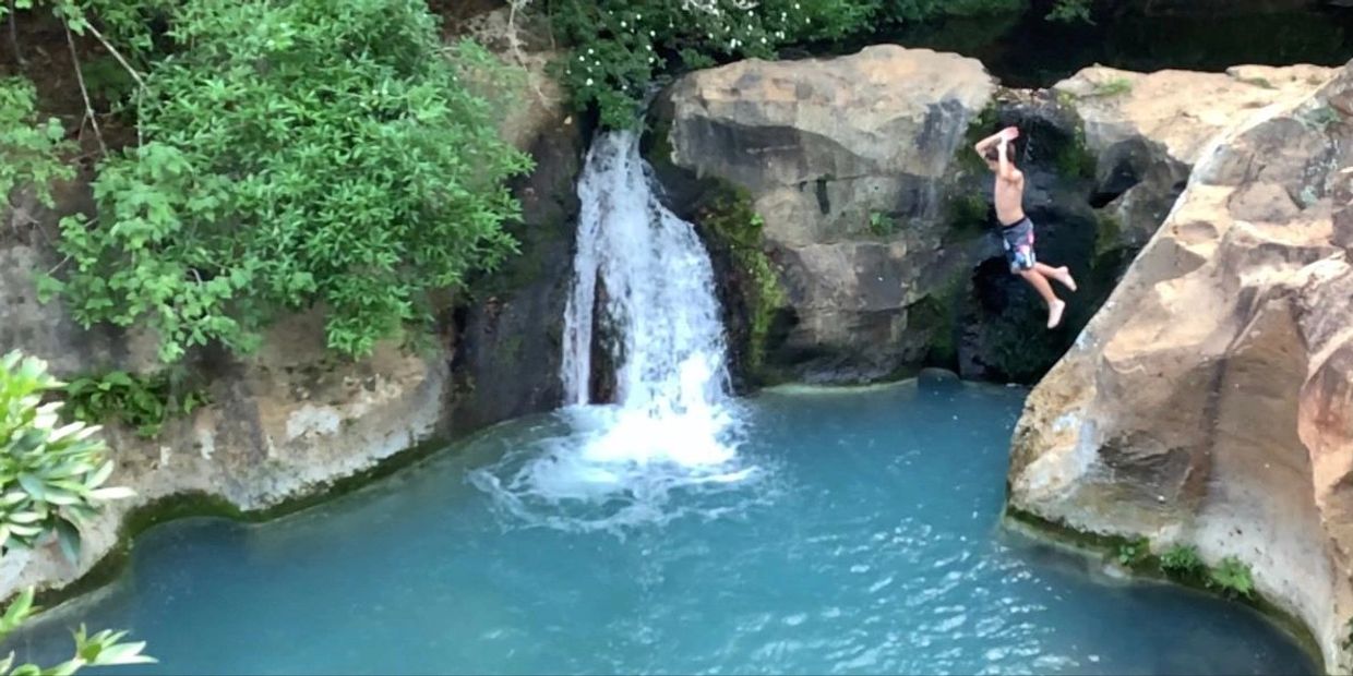 Boy jumping from a rocky cliff into a blue body of water with a waterfall in the background.