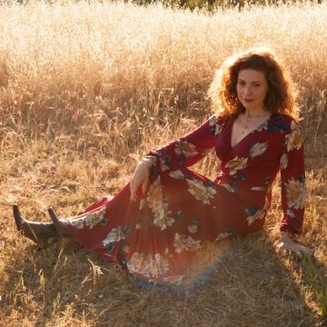 Susanna Brisk with red hair, sitting with legs outstretched in Topanga Canyon field in burgundy dres