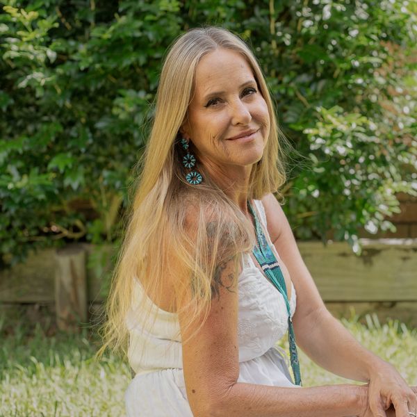 Smiling woman with long blonde hair outdoors in white dress and turquoise earrings.