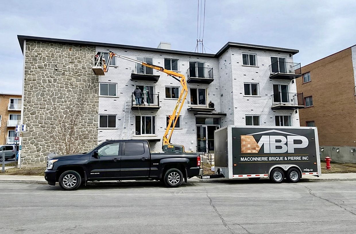 Workers using a lift to repair a building's exterior with a black truck and trailer parked nearby.