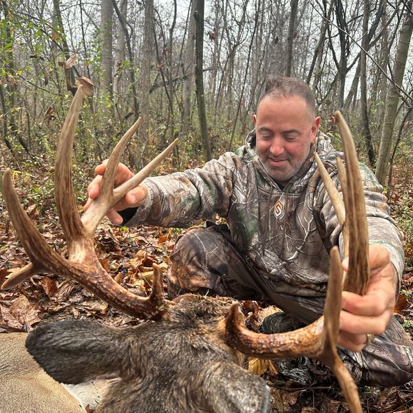 A hunter proudly holds up the antlers of a large buck in a forest.