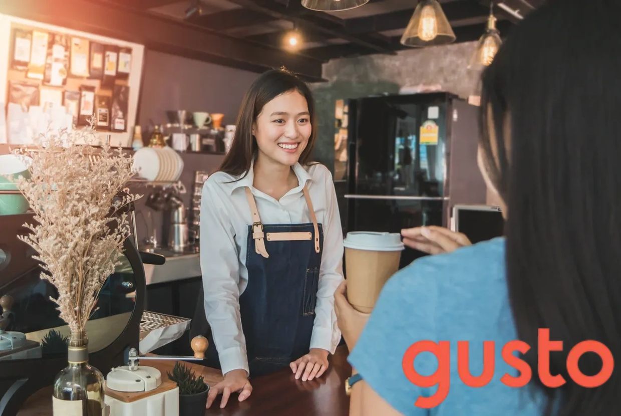 A smiling barista in a modern coffee shop.  'Gusto' is displayed-the payroll & HR services brand.