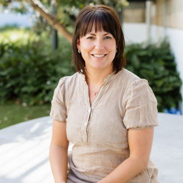 Smiling woman sitting outdoors with greenery in the background.