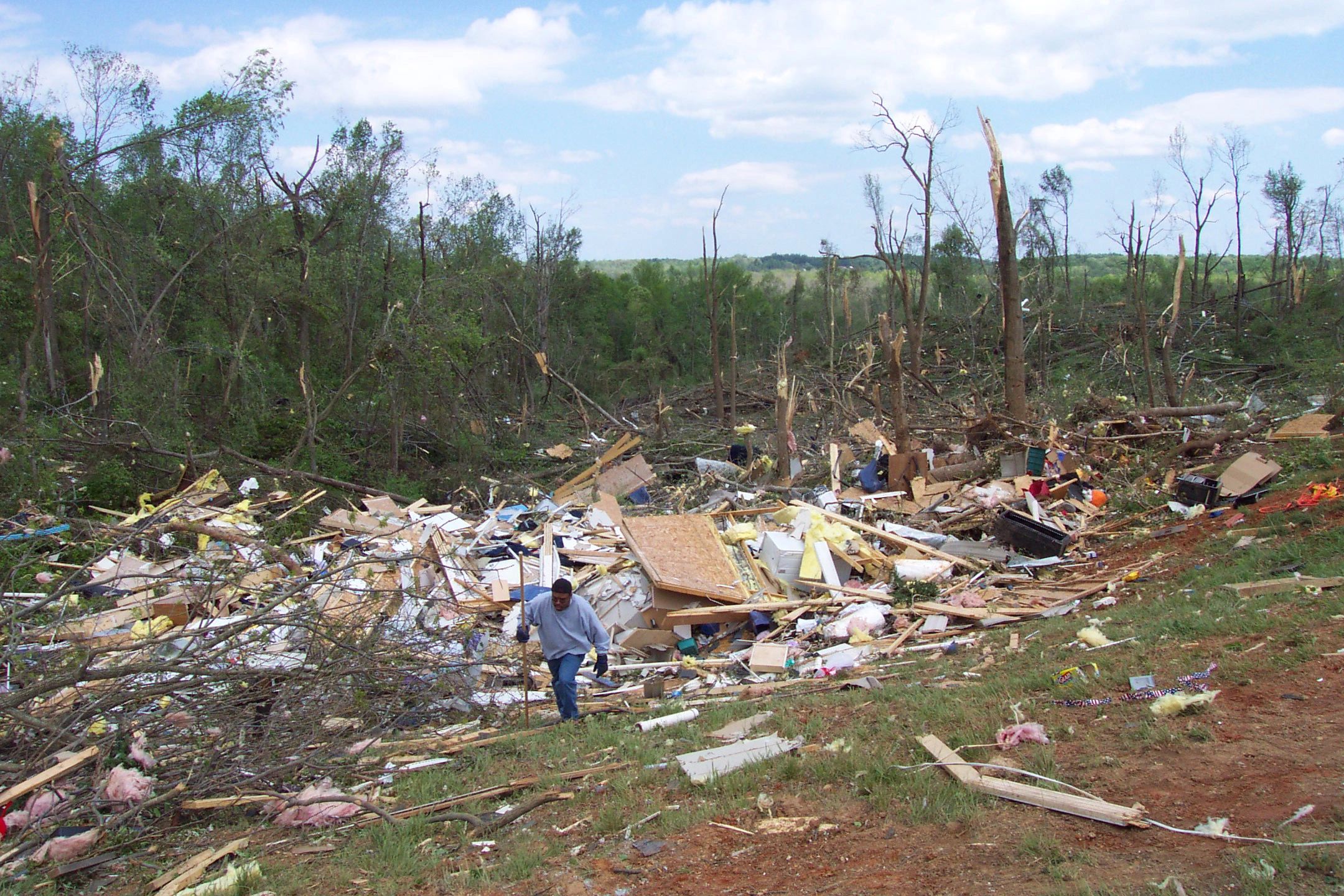 Man walks through debris of destroyed homes in wooded area after disaster.