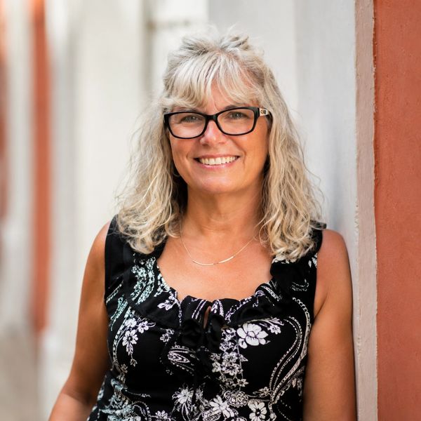 Smiling woman with glasses in a black floral dress leaning against a wall.