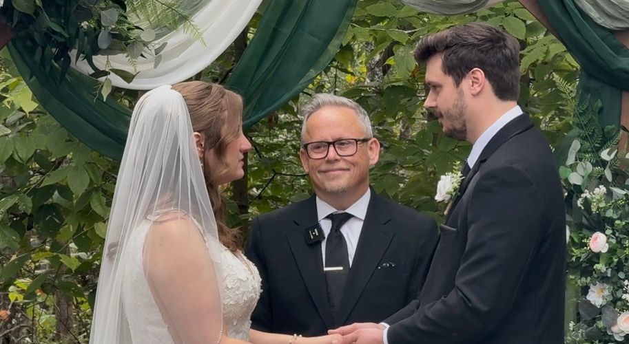 Bride and groom holding hands during wedding ceremony officiated by a man in glasses.