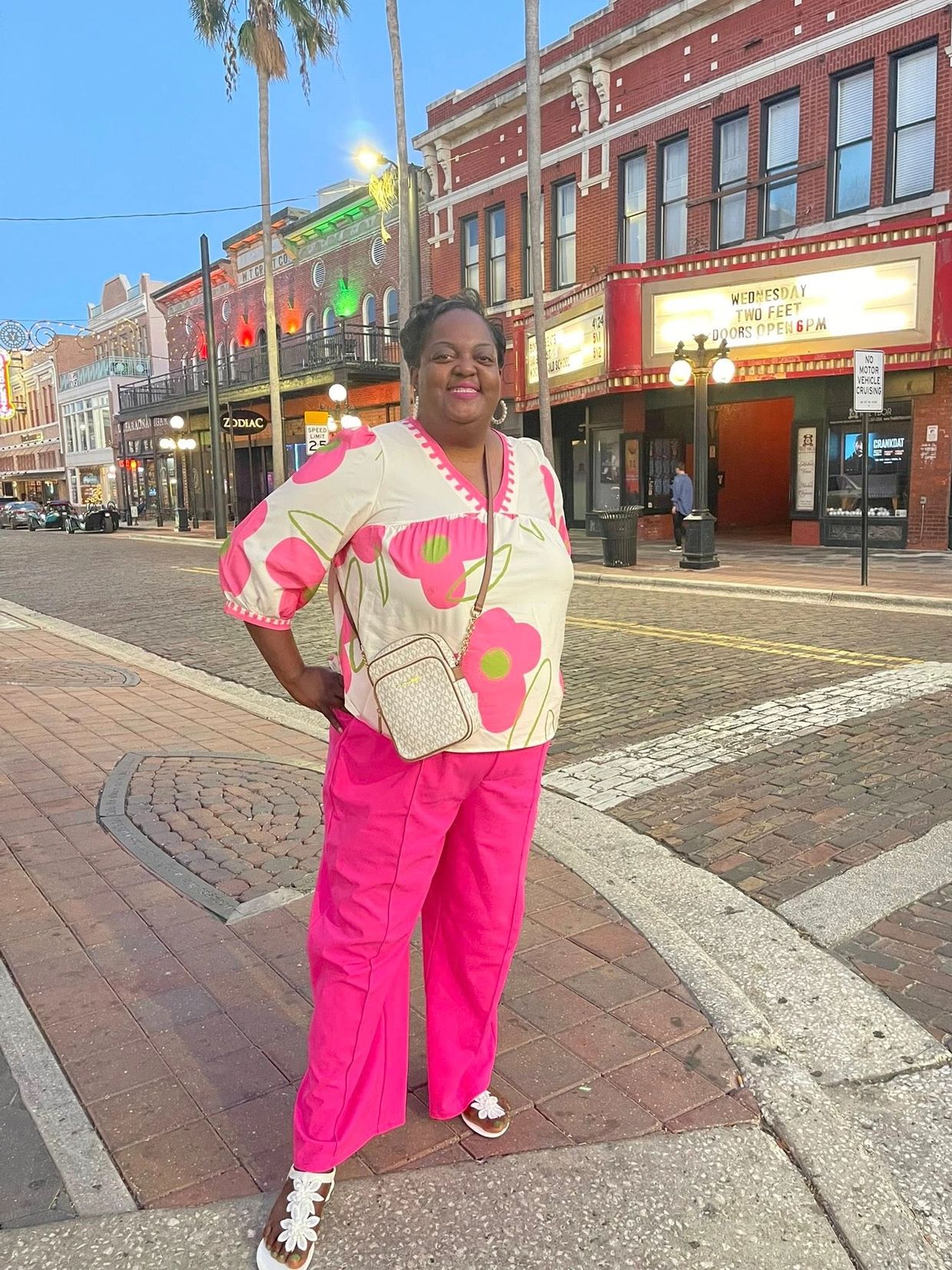 Smiling woman in bright pink pants and floral top stands on a brick-paved street in a lively downtown area.