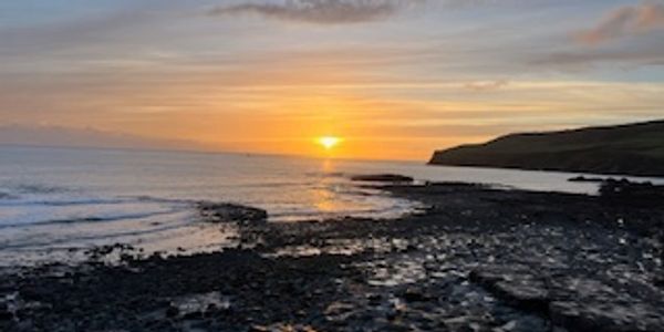 Sunset over a rocky shoreline with calm sea and distant hills.