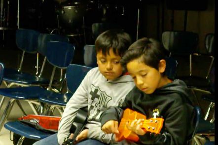 Two boys with medium skin and dark hair playing ukulele in a classroom.