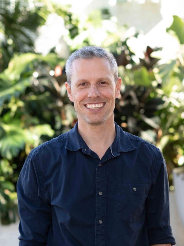 Smiling man in a navy blue shirt outdoors with greenery.