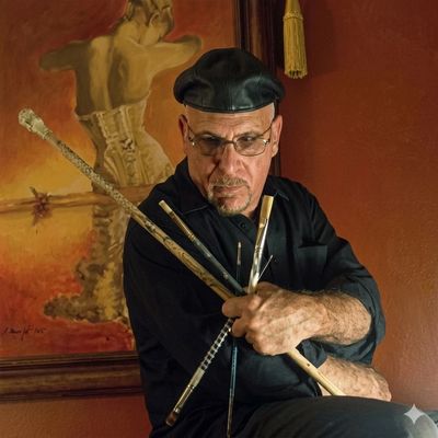 Man in a black beret poses with several flutes in front of a painting.
